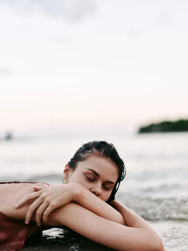 A young woman rests on a shallow shoreline, eyes closed, embraced by her arms as gentle waves lap the sand under a soft sunset glow.