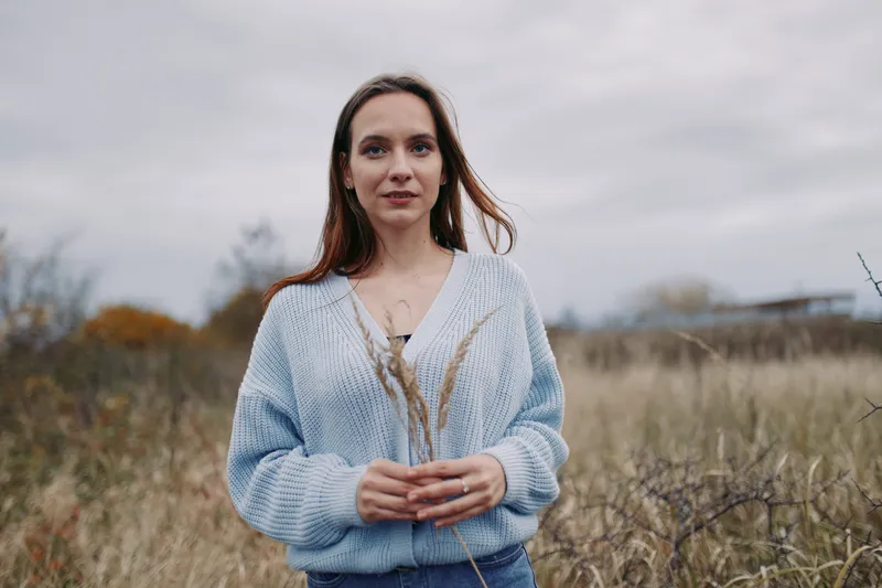 Young woman stands in a tall grassy field wearing a soft blue sweater, holding dried stems and looking at the camera in an unposed, candid portrait that conveys authenticity and credibility.