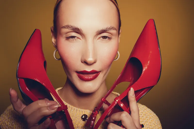 Close-up of a woman holding red high heels near her face with a playful expression. Warm lighting highlights her makeup and accessories on a yellow background.