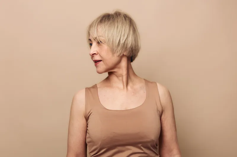 Portrait of middle aged woman with short blonde hair wearing beige tank top looking sideways on plain beige background. Concept of natural beauty, aging, and calm introspection in studio.