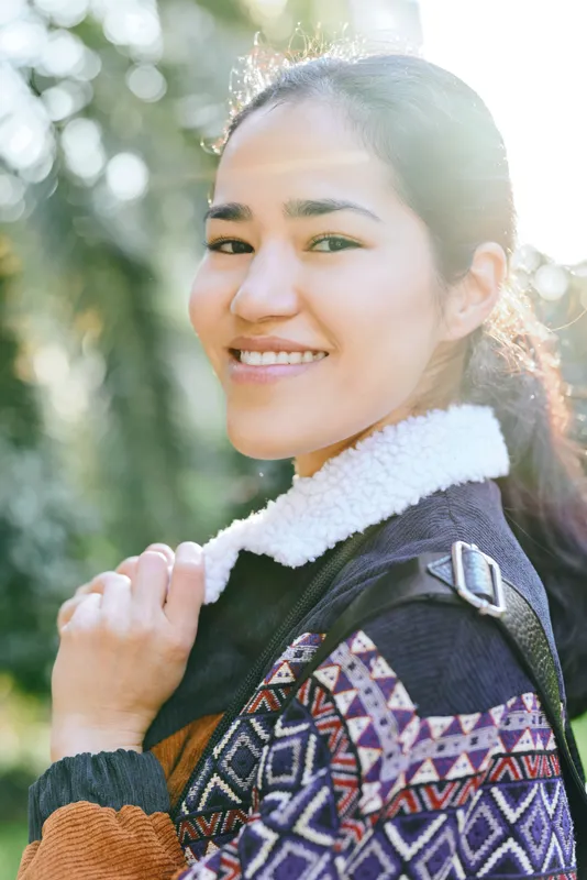 Woman wearing a jacket outdoors with a smile, patterned fashion, cozy textures and sunlight in a leafy park setting, showcasing casual mood and sunny ambience