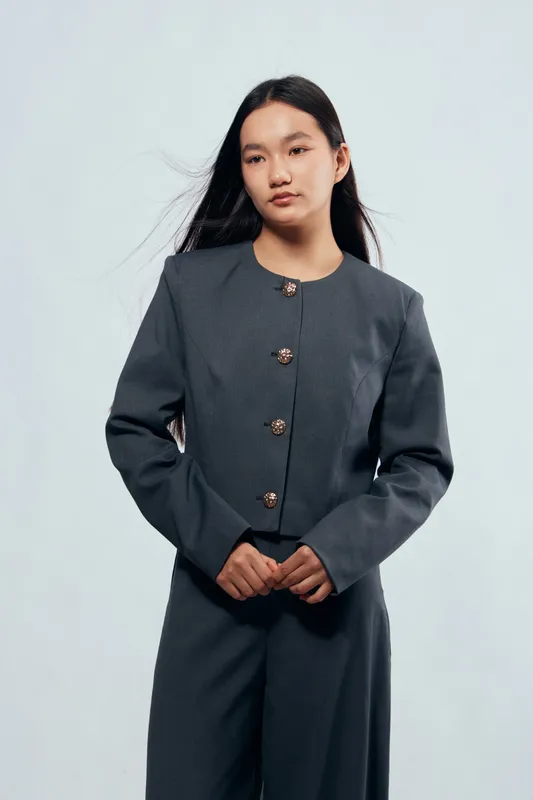 Young woman with long dark hair wearing gray suit jacket and pants standing against plain background with serious expression and hands clasped for fashion or business portrait.