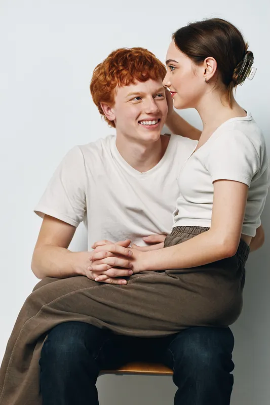 Young couple sitting together with affectionate smile and gentle embrace on white studio background. Love, relationship, casual clothing, happiness and intimacy.