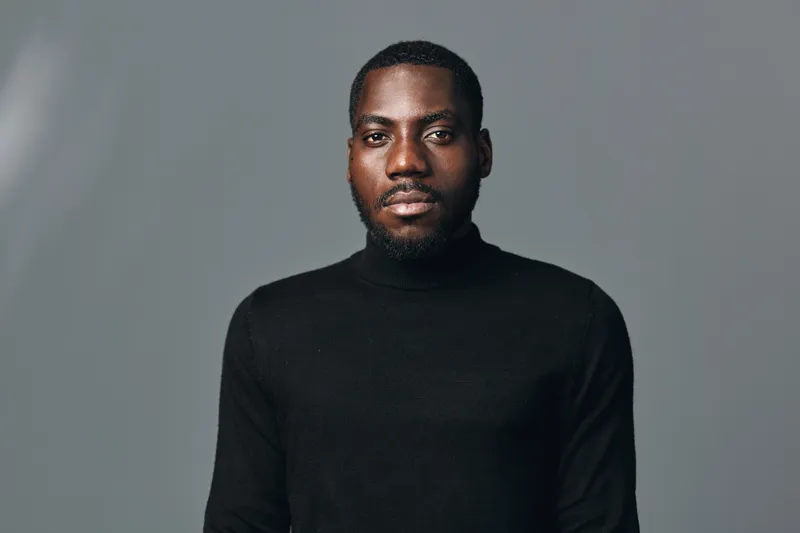 African man portrait with serious expression wearing black turtleneck sweater in creative conceptual visual style on gray background. Studio shot with focused lighting and neutral tone.