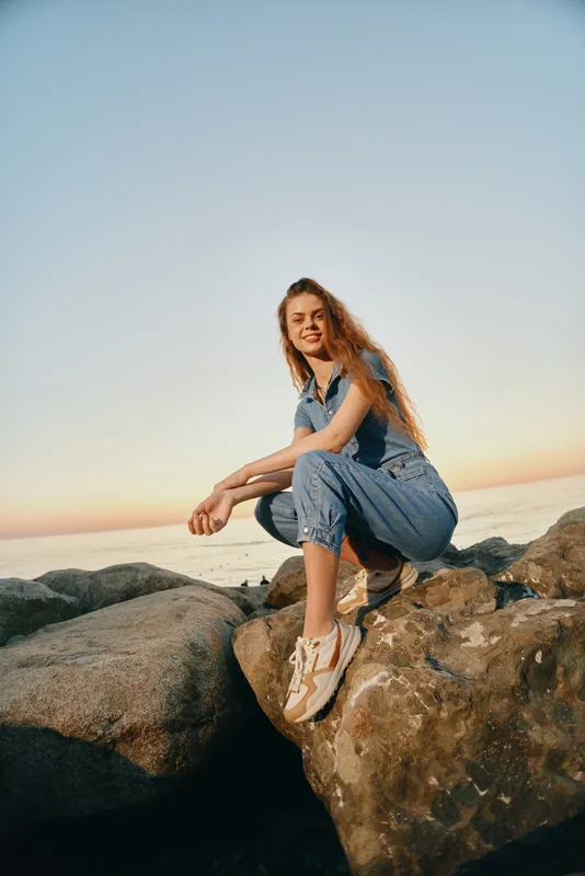 Lifestyle woman in modern boho-western denim street style sitting on rocks by the sea during golden hour with warm film color tones and relaxed expression