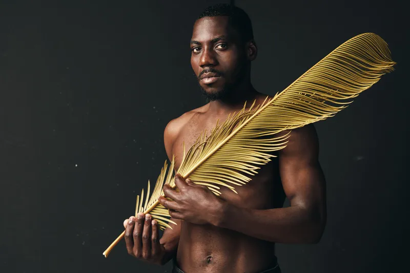 Black man with dark skin holding large golden feather in creative visual design style. Artistic portrait with bare torso and serious expression on dark background for concept.