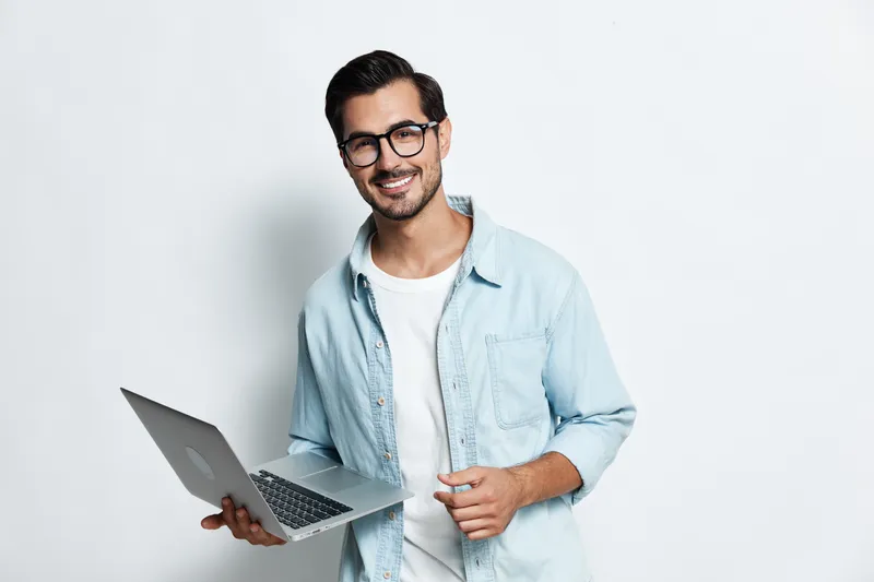 Smiling young man wearing glasses holds laptop, looking at the camera with a friendly expression against a solid light background. Professional lifestyle concept.