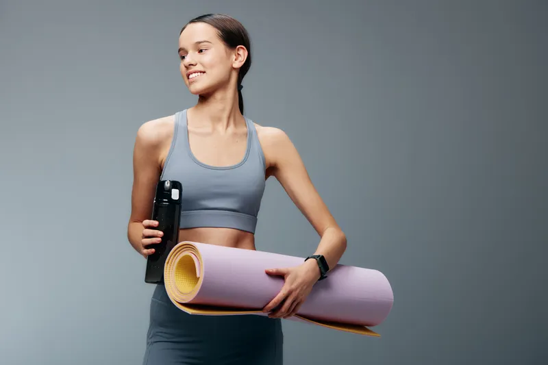 Young woman in athletic wear holds a water bottle and a rolled yoga mat, ready for training. Minimal studio backdrop highlights fitness focus and personal health goals.
