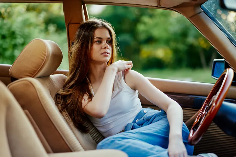 Young woman relaxing in a vintage car, wearing casual clothing, with natural background, conveying a sense of freedom and nostalgia, ideal for summer themes.