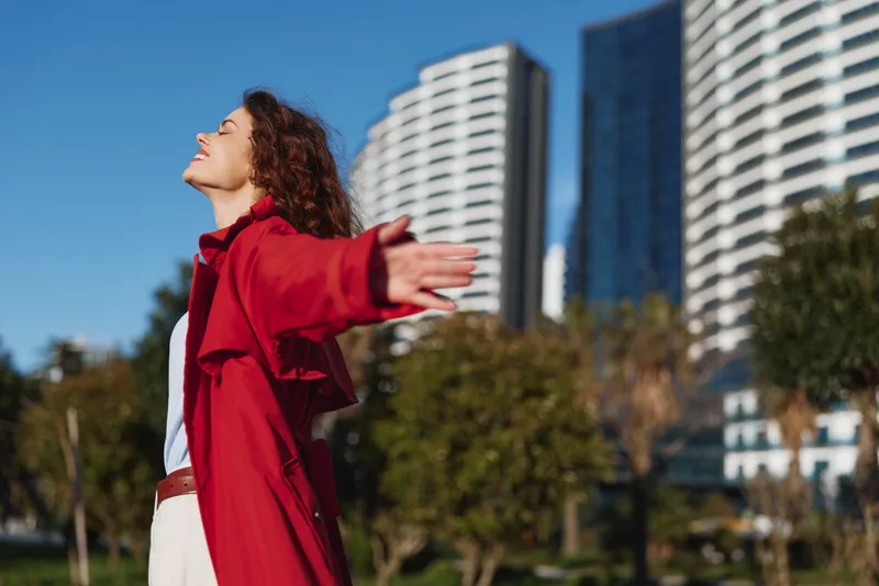 Young woman in red coat enjoys fresh air with open arms in urban park on sunny day with modern buildings in background and clear blue sky.