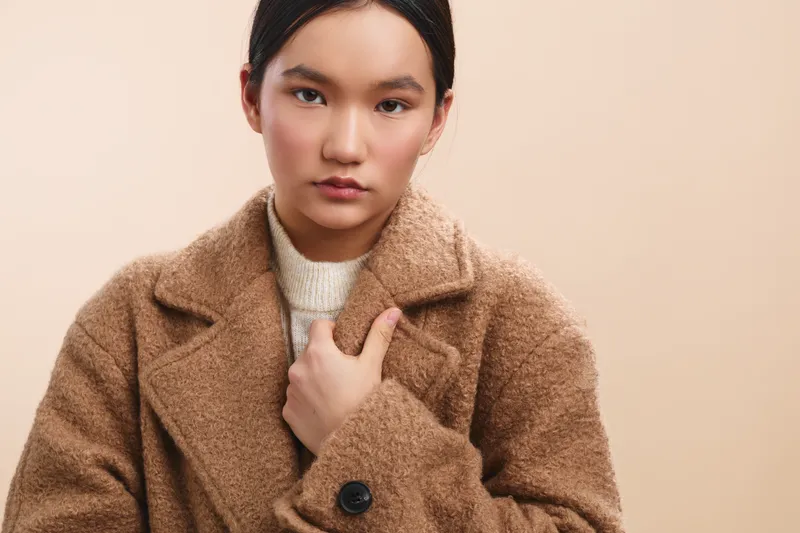 Asian woman wears a warm beige coat against a neutral studio backdrop, creating a timeless portrait that emphasizes calm confidence and minimalist fashion.