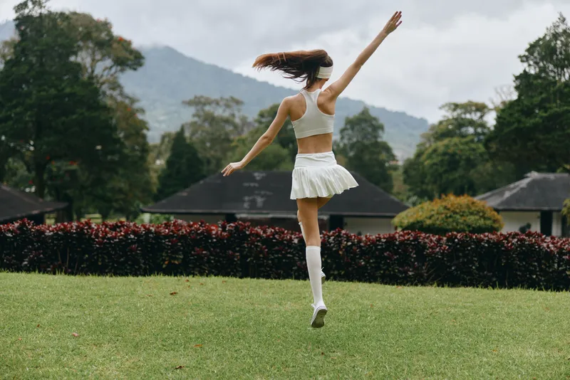 Young woman in sporty attire joyfully jumping on a green lawn, wearing a white crop top and skirt, embodying happiness and vitality in a vibrant outdoor setting