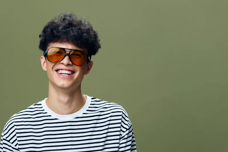 smiling young man in striped shirt and oversized sunglasses with curly hair on green background expressing happiness and casual style with confident attitude