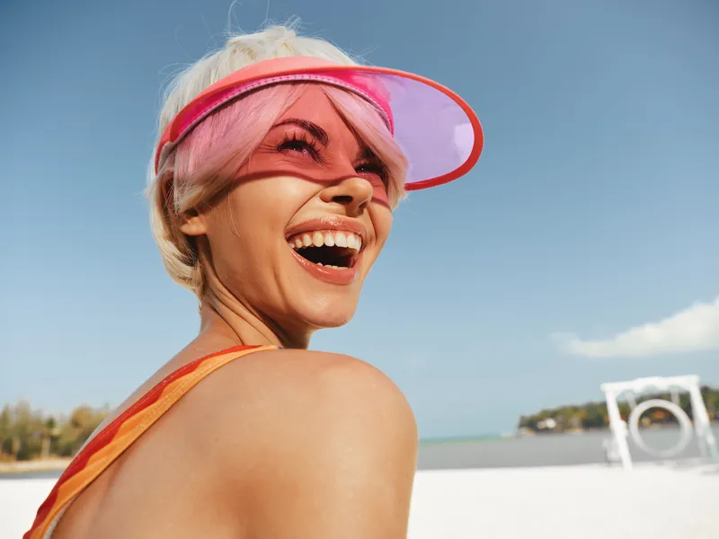 Bright sunlit portrait of a joyful woman wearing a pink visor on a sunny beach, captured from behind as she laughs, with clear blue sky and white sand behind her.