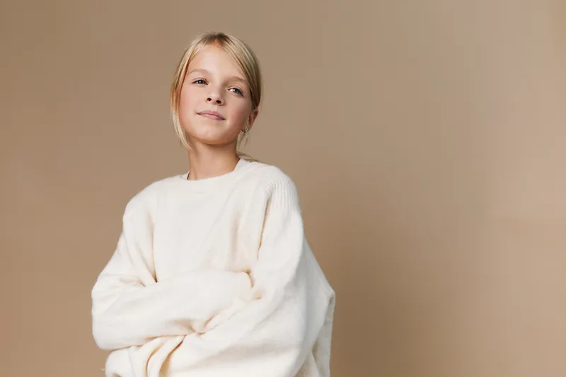 Young girl in an oversized white sweater stands against a warm beige backdrop. Soft lighting highlights her relaxed pose and confident, gentle expression.
