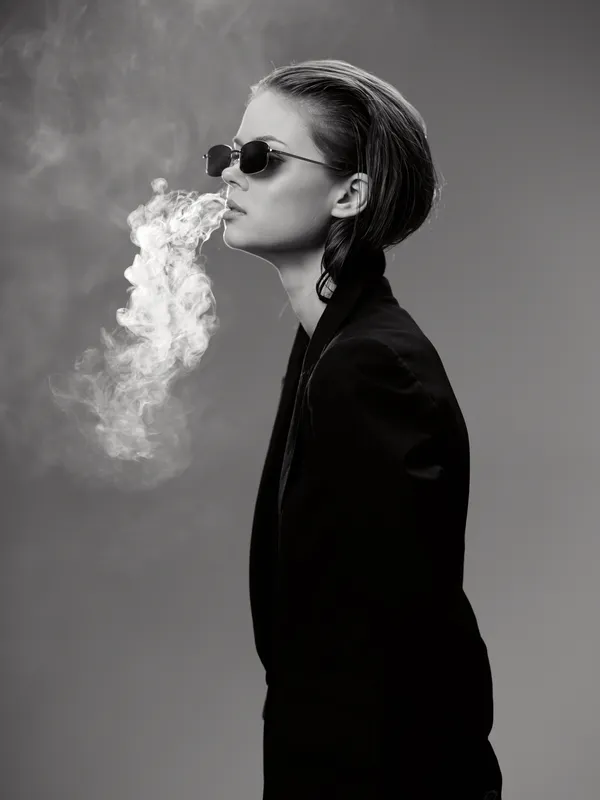 Black and white portrait of a confident woman exhaling smoke in stylish sunglasses and dark clothing