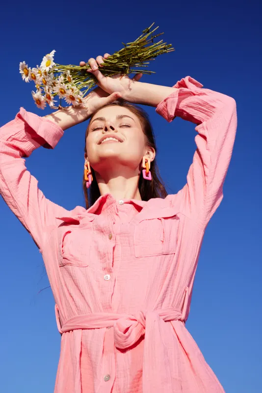 Young model in a pink outfit holding flowers against a clear blue sky, expressing joy and tranquility, perfect for spring themes and fashion imagery