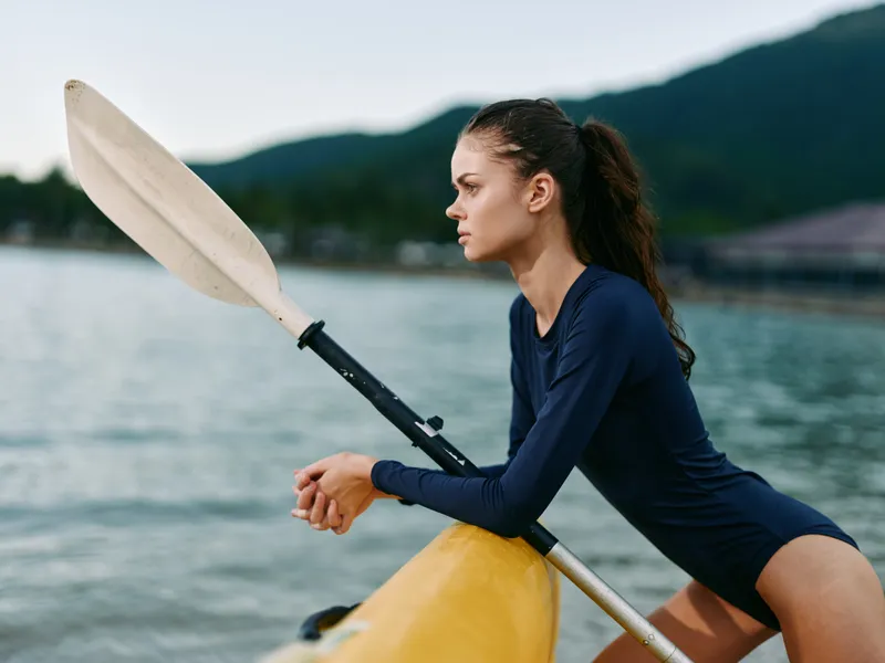focused woman kayaking on calm water with paddle resting, wearing navy long sleeve shirt, outdoors with mountains background