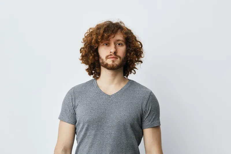 Confident young man with curly hair in a gray t shirt against a light background, expressing calmness and serenity, ideal for lifestyle and wellness themes