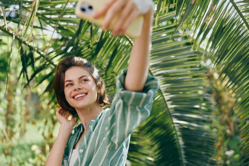 Portrait of a smiling woman taking a sunny selfie outdoors among palm leaves, wearing a striped shirt, capturing a carefree tropical moment.