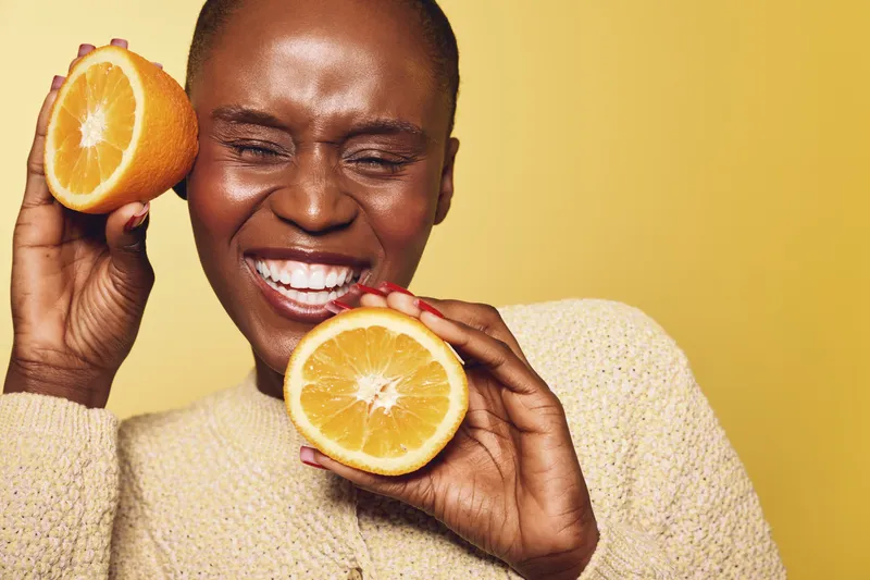 Happy African woman with dark skin holding two orange halves near her face, smiling with closed eyes. Studio shot on yellow background. Concept of healthy lifestyle and natural beauty.
