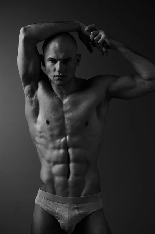 Black and white image of a muscular man posing confidently, showcasing his toned physique and strength. Strong lighting emphasizes the contours and shadows on his body.