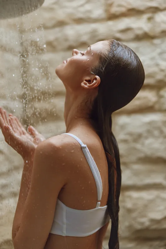 young woman enjoying natural shower, wet hair, relaxed pose, warm textured stone wall background, peaceful outdoor moment, summer wellness