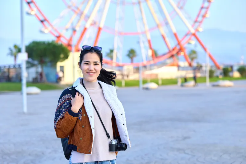 Woman with camera in outdoor amusement park, smiling and casual, carrying a bag, ferris wheel in the background, sunny day, travel vibe, candid street photography, lively and natural scene