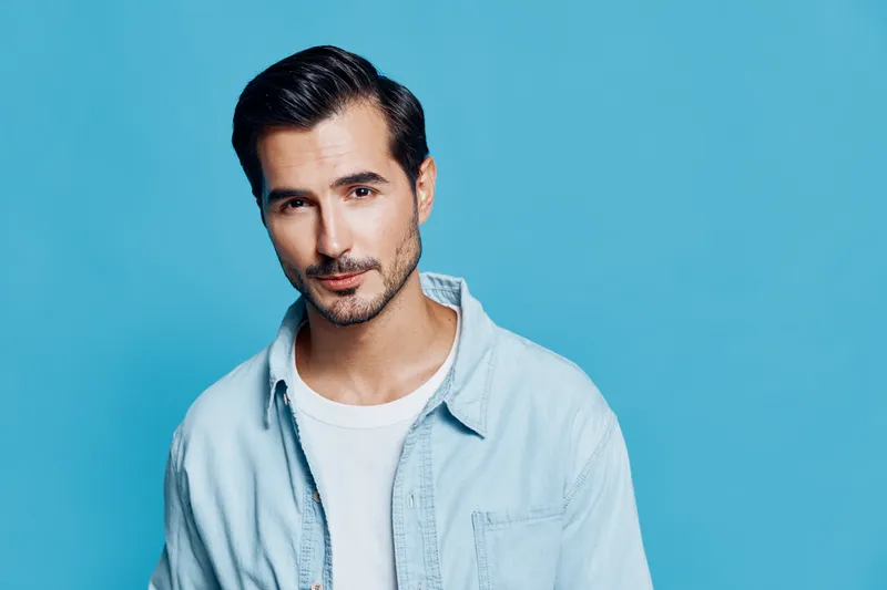 Handsome smiling man wearing light blue shirt and white t-shirt, looking confidently at the camera against a solid turquoise background. Lifestyle concept.
