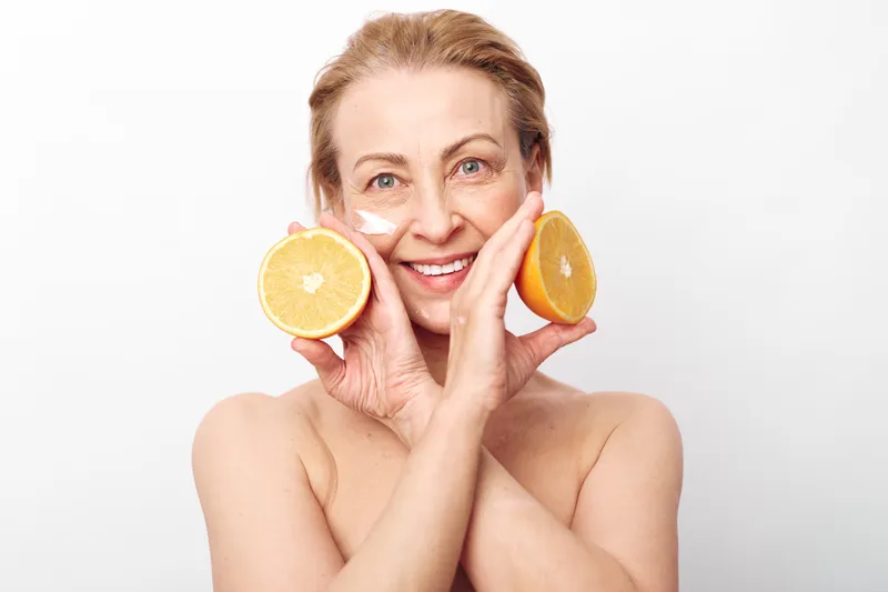 Smiling middle aged woman with natural skin applies white cream on cheeks holding fresh orange halves near face on white background. Concept of skincare, healthy lifestyle, and natural beauty.