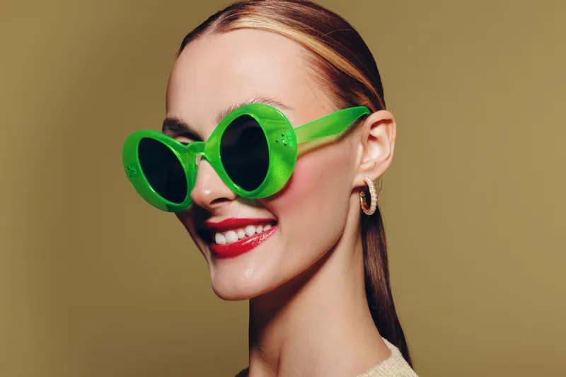 Smiling woman with slicked hair wears large green sunglasses and red lipstick. Studio portrait on beige background. Concept of fashion, style, and confident beauty.