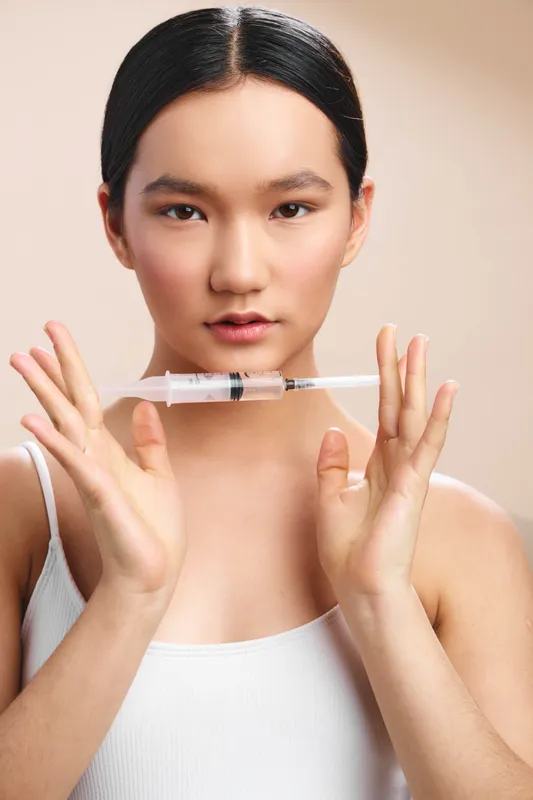 asian woman presents a closeup in a clinical setting, holding a syringe near her face with a calm, confident expression, conveying professional healthcare atmosphere.