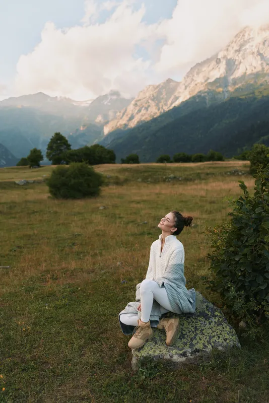 A young woman in a white dress kneels on a rock in a sunlit meadow with mountains rising behind her, looking upward with a serene contemplative smile