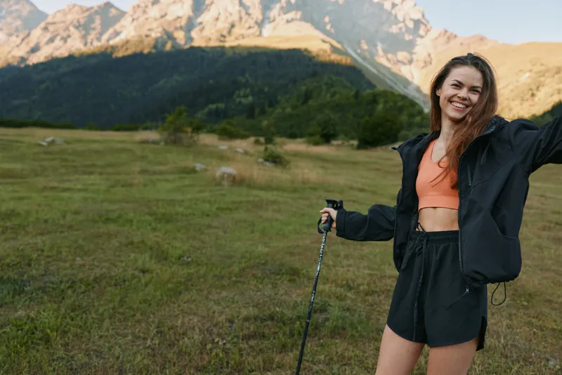 A cheerful woman hikes through a bright meadow beneath towering mountains, enjoying a sunny day and fresh air, embodying outdoor adventure and wellness.