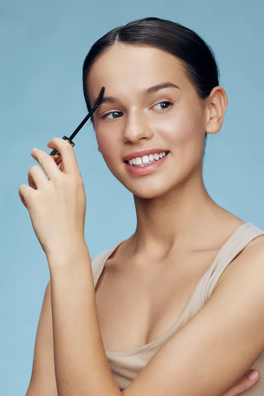 Young model applying eyebrow gel with a joyful expression, showcasing natural beauty and makeup preparation in a soft blue background, perfect for beauty and cosmetics themes