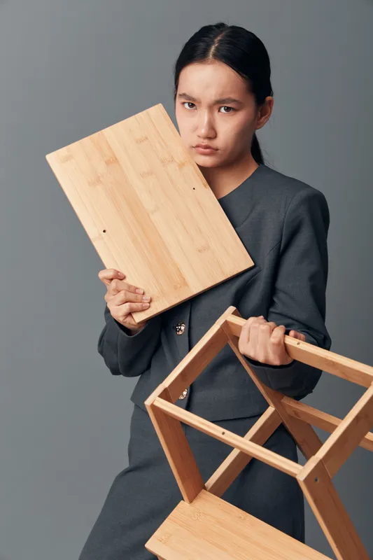 Young woman assembling wooden furniture parts indoor holding wooden board and frame in hands, focused girl repairing or building with tools on gray background.
