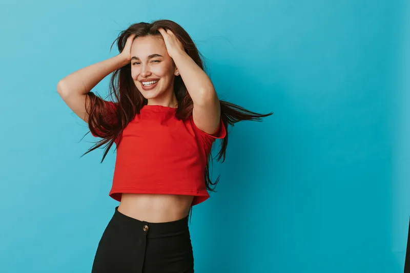 Young woman smiling with hands in hair wearing red crop top and black pants standing against blue background. Happy female model posing with long hair and casual outfit in studio.