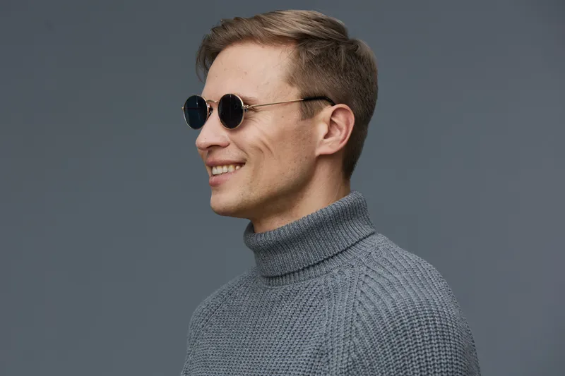 Stylish young man with light brown hair wearing black round sunglasses and grey turtleneck sweater smiling against a plain grey background. Casual fashion portrait with a modern, cheerful vibe. People