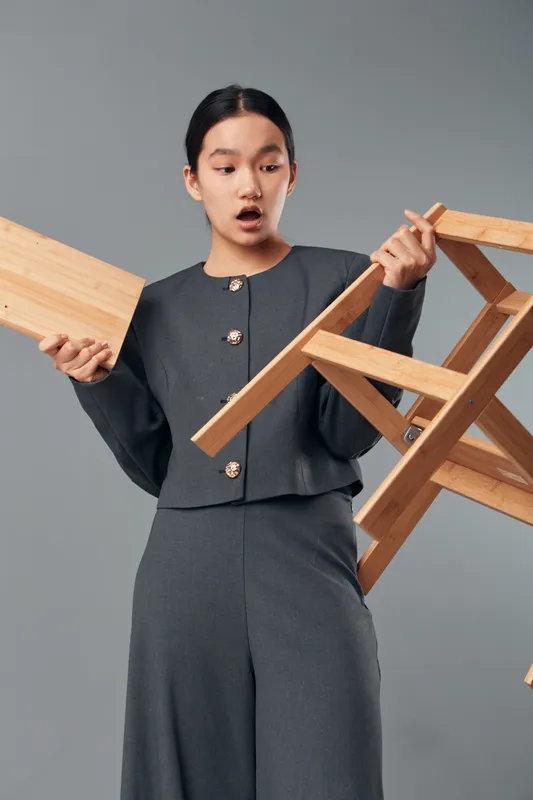 Woman with confused expression holding broken wooden chair and detached piece in studio, casual outfit, isolated on gray. Repair, furniture, problem, hands, chaos, damage, indoors.