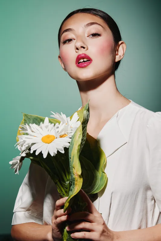 Young woman with shiny red lipstick holding white daisy flowers wrapped in large green leaves on green background. Stylish beauty fashion and creative colorful concept.
