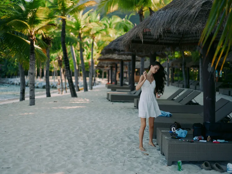 A woman in a flowing white dress stands on a sandy beach among palm trees and thatched huts, capturing a serene vacation moment with graceful posture and sunshine.
