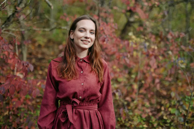 A candid portrait of a woman standing amid autumn foliage, wearing a burgundy dress. Natural lighting conveys sincerity and genuine mood.