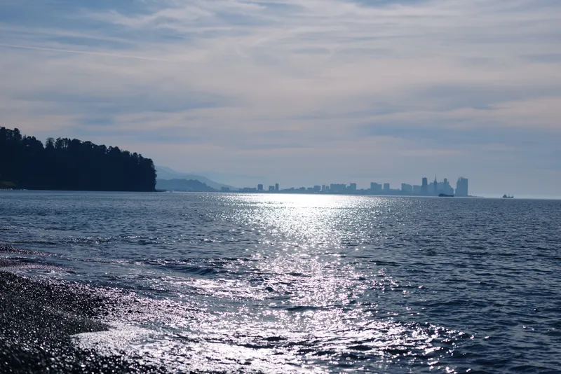Ocean and distant city skyline across calm water, sunlight glittering on waves, rocky shore in the foreground, tranquil seascape under a pale blue sky.