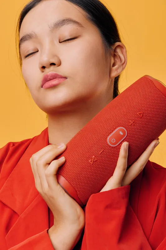 Young woman holding red wireless speaker close to face with closed eyes, enjoying music, wearing red jacket. Studio portrait on yellow background, relaxed and peaceful mood.