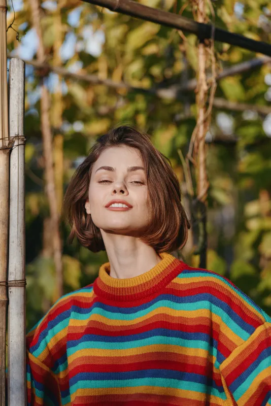 Young woman in a colorful striped sweater poses in an outdoor portrait, short bob hair, smiling with closed eyes while leaning on a metal post in an autumn garden.
