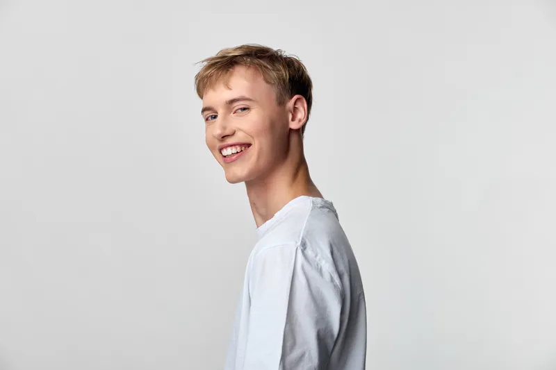 Smiling man in a light blue shirt stands in side profile against a neutral backdrop, exuding warmth and relaxed confidence for lifestyle, fashion, and wellness contexts with a simple contemporary