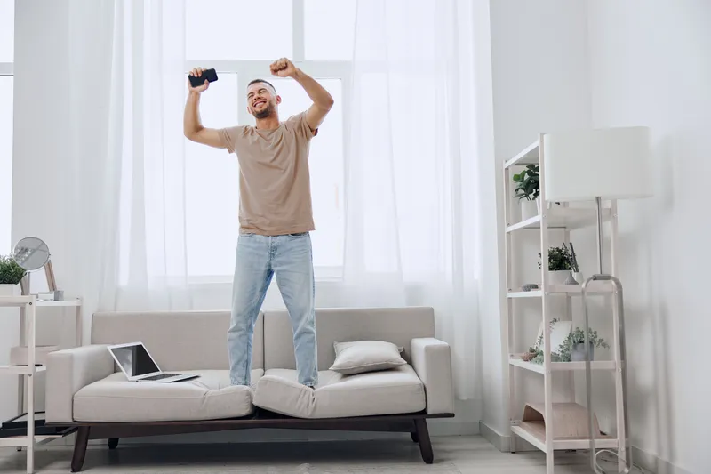 Joyful man celebrating success while standing on a couch, expressing excitement and overcoming challenges in a bright, minimalist living space.