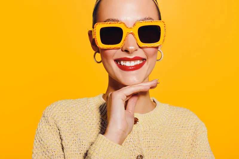 Smiling woman with bright red lipstick wearing large yellow sunglasses and beige sweater poses against vibrant yellow background. Concept of fashion, style, confidence, and summer mood.