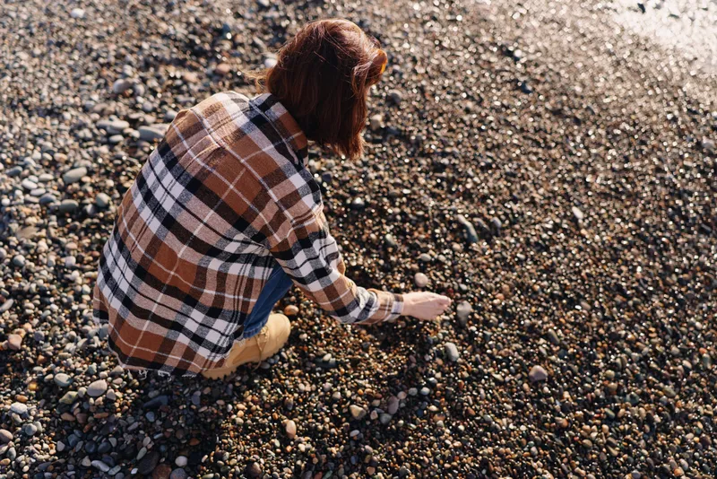 Person crouching on a rocky shoreline, examining pebbles with focus, wearing a plaid shirt, outdoor exploration and mineral collection under warm sunlight.