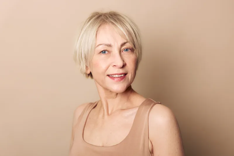 Portrait of a middle aged woman with short blonde hair smiling gently wearing a beige tank top against a beige background. Concept of natural beauty, confidence, and mature skin care.